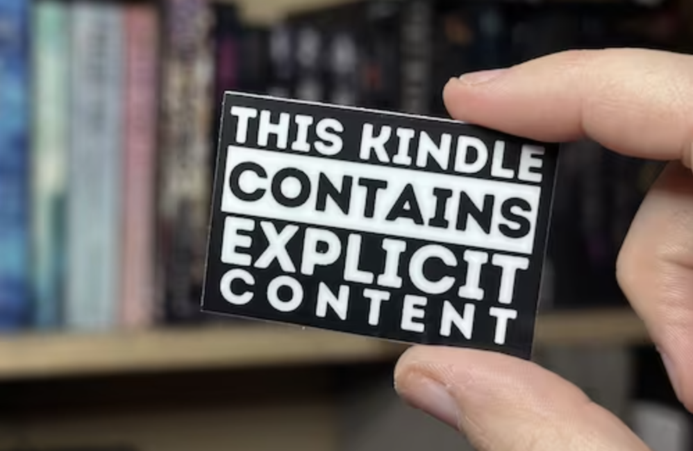 A black and white pin that reads "this Kindle contains explicit content" held in front of bookshelves