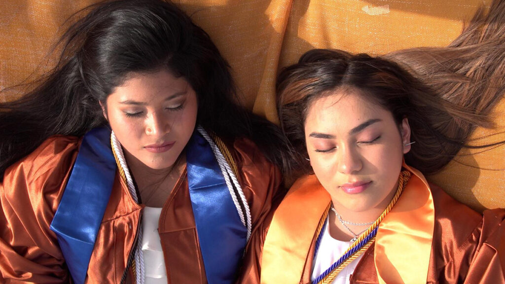 SXSW 24 film "The In Between" Paola and Marianne in a scene from The In Between. visual description: Two young women lying with their eyes closed on an orange mat in burnt orange graduation robes. | Credit: Robie Flores
