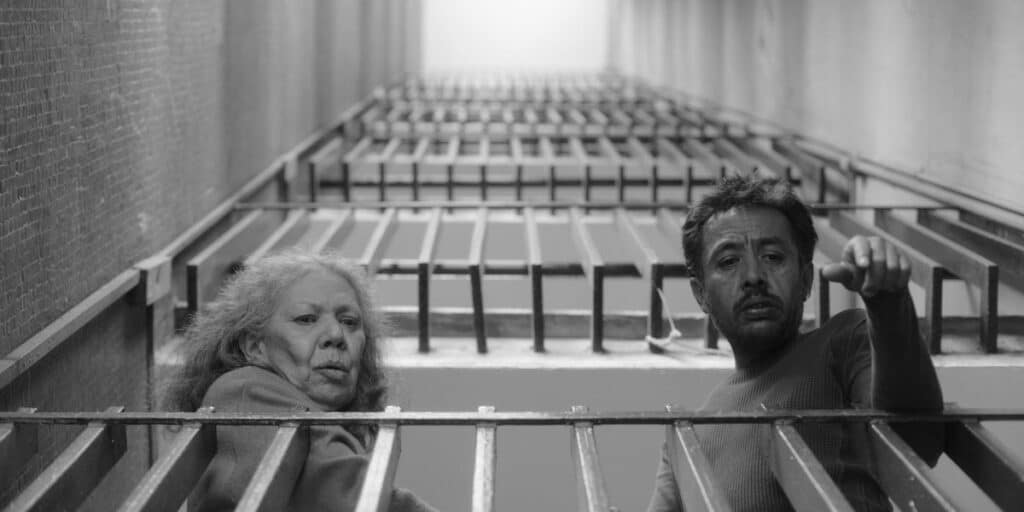 A black-and-white still from "We Shall Not Be Moved" shows an older woman and a middle-aged man positioned behind a set of metal bars in a confined, industrial-looking corridor. The man gestures forward while both figures look off-frame. The repeating bars and tight space emphasize confinement and tension within the scene.