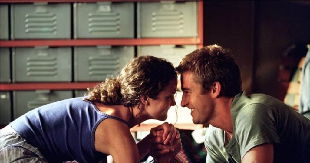 A still from a television drama show, "Felicity." Two young adults seated close together, leaning forward with their foreheads nearly touching as they smile at one another. Their hands are clasped between them, suggesting intimacy and trust. The setting appears informal and private, with muted lighting and shelves lined with storage bins in the background. The image captures a quiet, emotionally charged moment that emphasizes connection, vulnerability, and the tenderness of young adulthood.